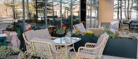 An outdoor seating area with wicker furniture and planters, large windows, and a view of trees and buildings.