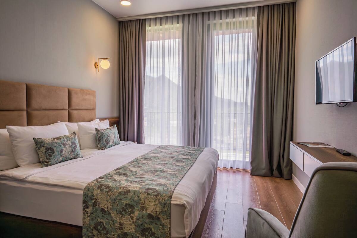 A hotel room with a large bed, a desk with a flat-screen TV, and a view of mountains through sheer curtains.