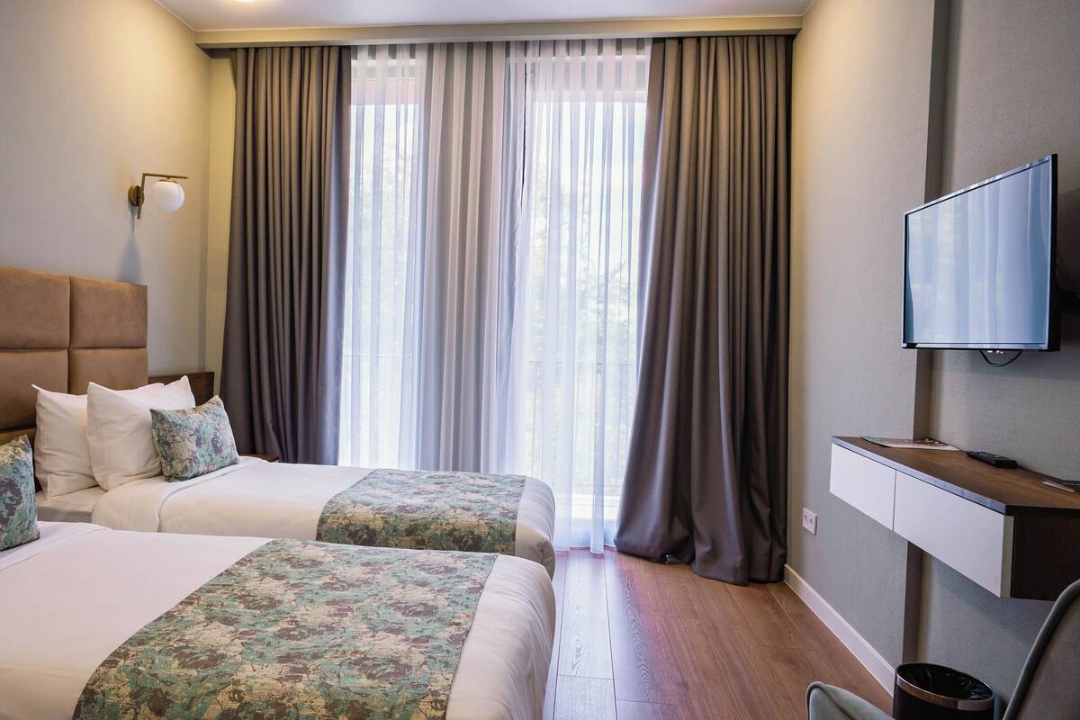 A hotel room with two beds, a large window with curtains, a mounted TV, and a wooden floor.