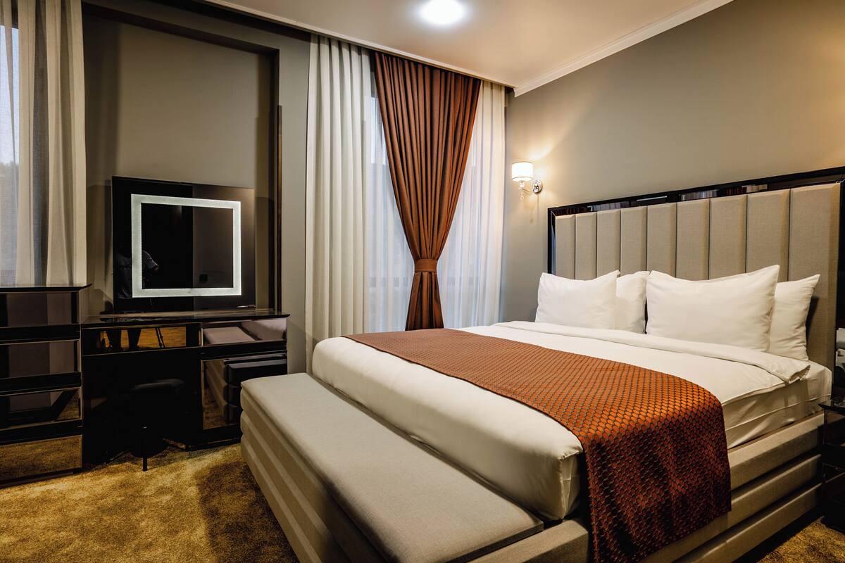A hotel room with a bed, a TV, a desk, and a large window with curtains.