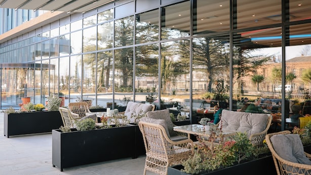 Un espacio al aire libre con mobiliario de mimbre y macetas, amplias ventanas que ofrecen vistas a la calle y a los árboles.