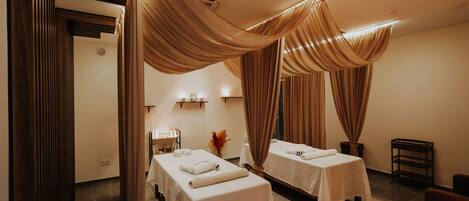 Un cuarto de spa con dos camas, cada una cubierta con una sábana blanca y una toalla, cortinas colgantes y una pequeña mesa con un florero.