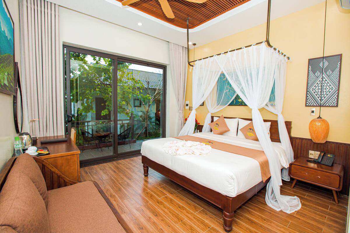 A spacious bedroom with a large bed, wooden flooring, a balcony with a glass door, and a view of greenery.