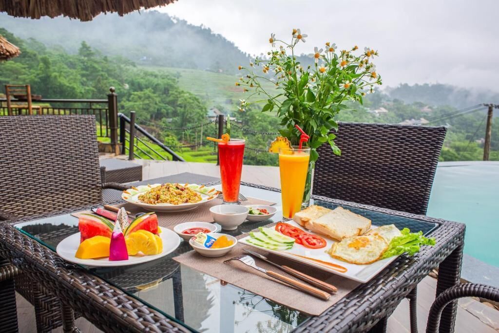 A table set with various dishes, drinks, and a vase of flowers, overlooking a lush green landscape.