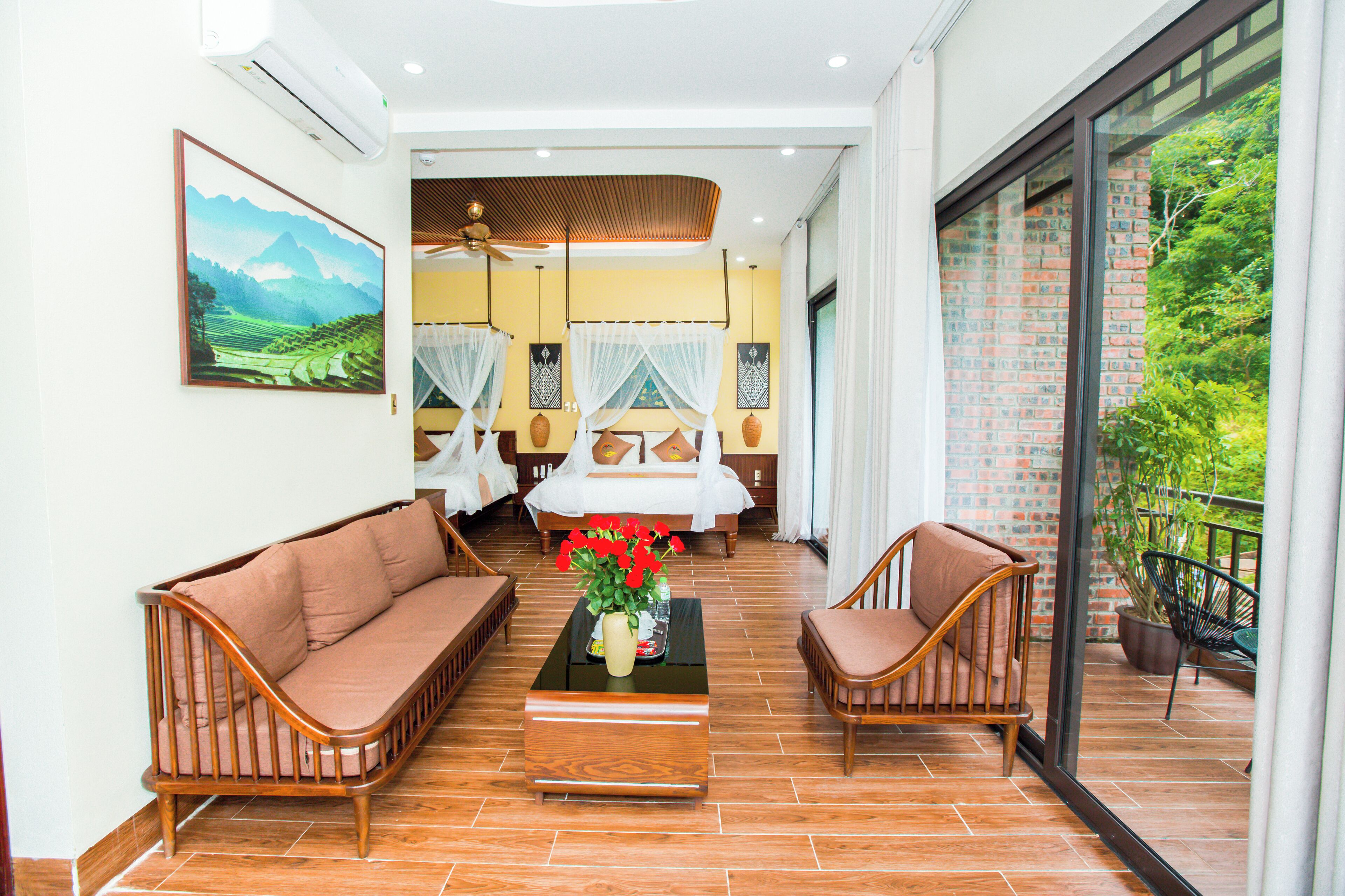 Amplia habitación con una cama grande, dos sofás, una mesa de centro con flores y un balcón con vistas a zonas verdes.