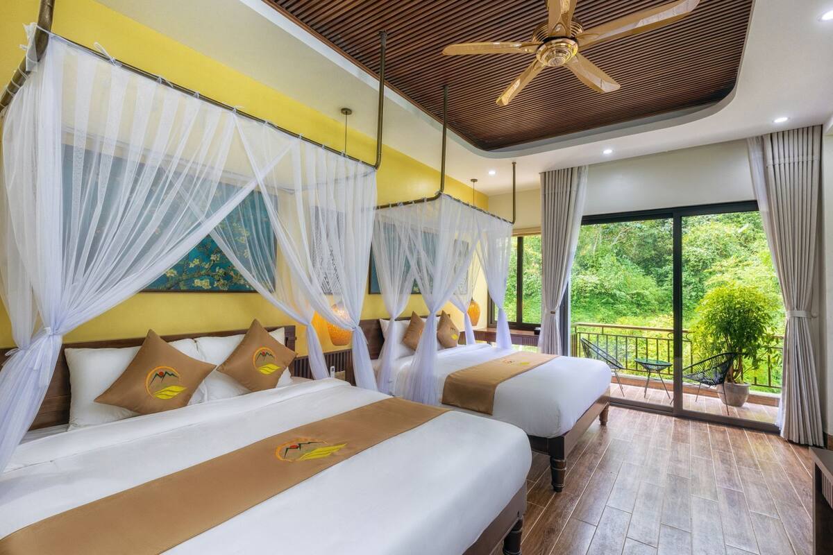 A hotel room with two beds, a ceiling fan, and a balcony with a view of greenery.