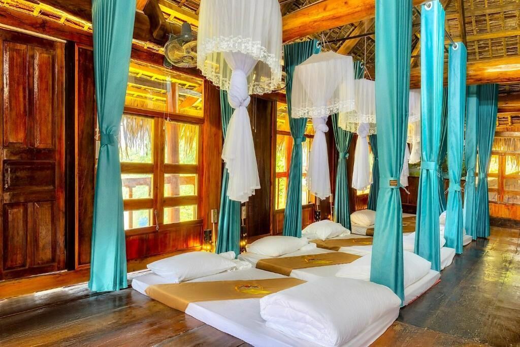 Una habitación con pisos de madera, cortinas azul verdosas y camas con dosel blanco suspendido.