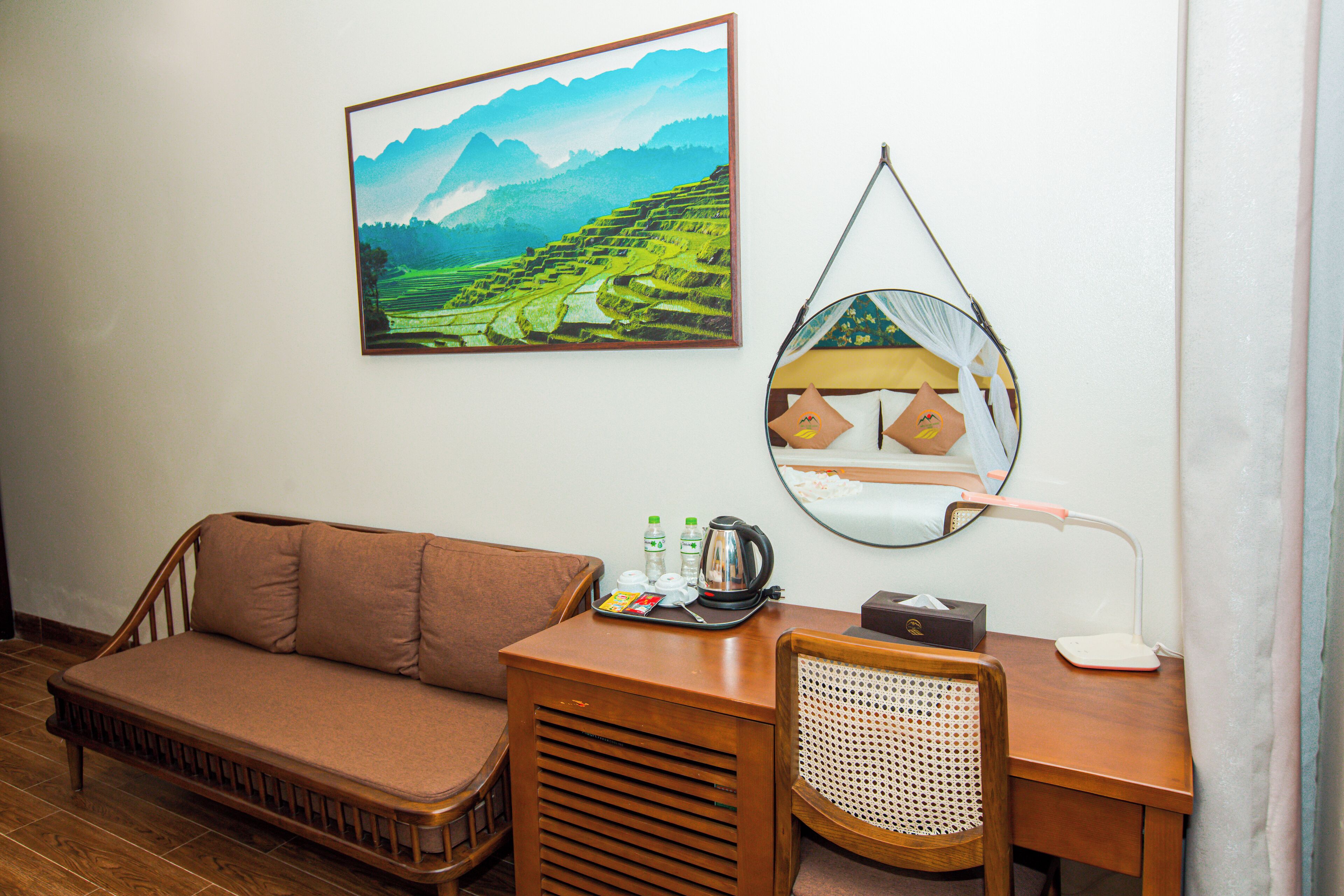 A room with a wooden sofa, a desk with a mirror, a framed landscape painting, and a tray with toiletries.