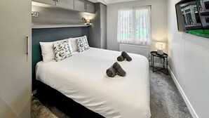 Ein modernes Hotelzimmer mit Bett, Nachttisch, Vorhängen am Fenster und einem an der Wand montierten Fernseher.