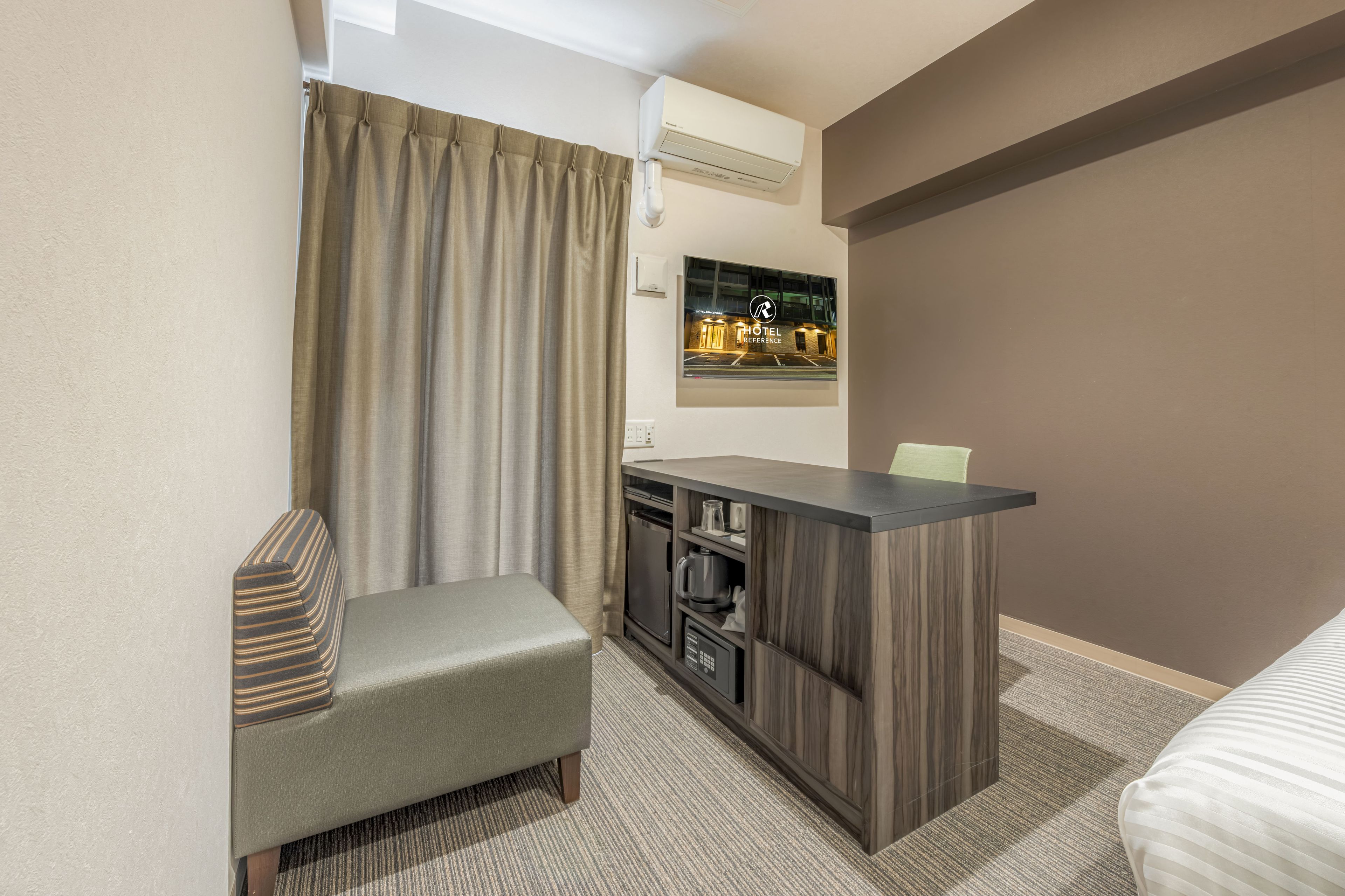 A hotel room with a bed, a small desk with a chair, a window with curtains, an air conditioning unit, and a framed picture on the wall.