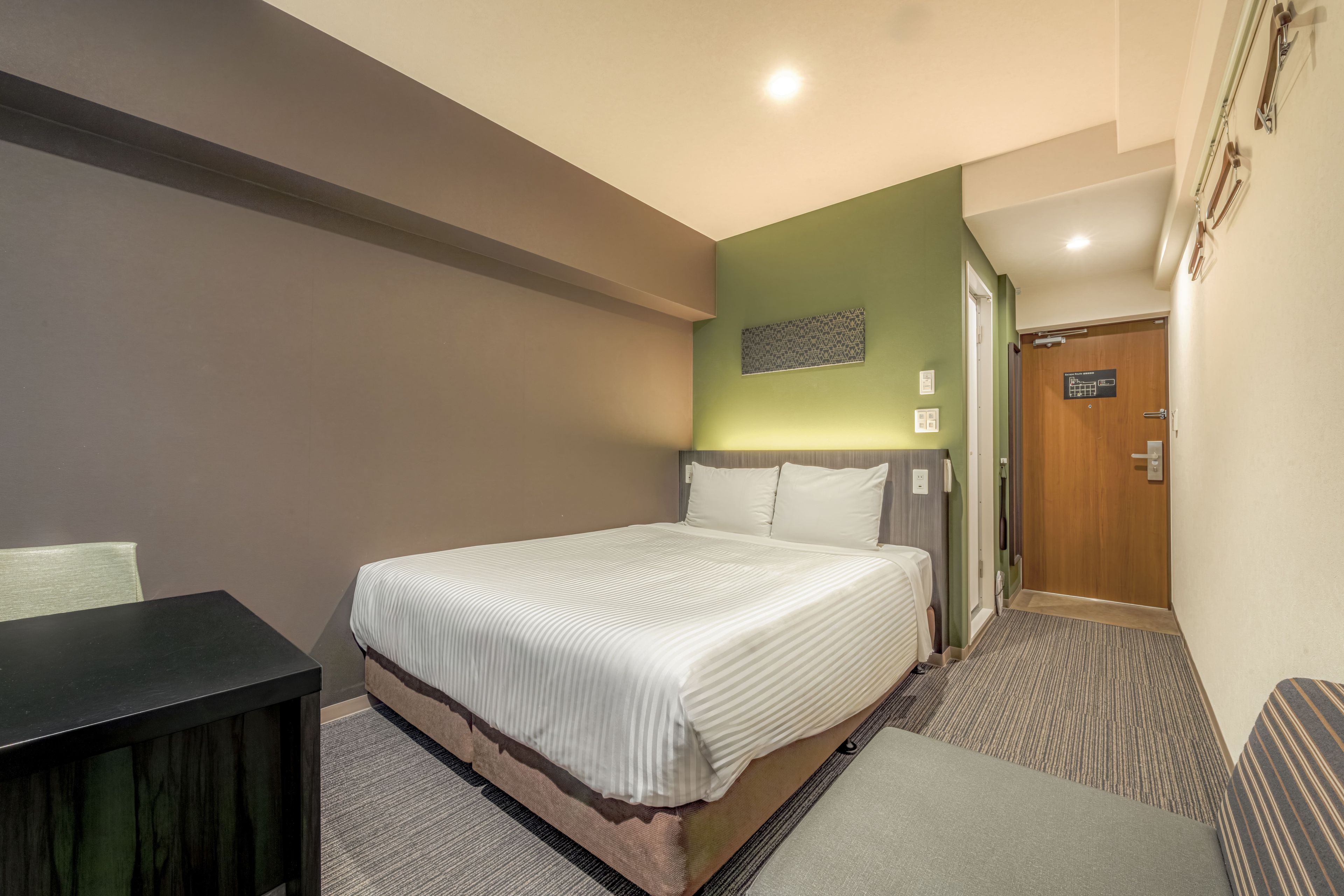 A hotel room with a bed, a desk, a door, and a wall-mounted light fixture.