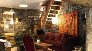 Un salon chaleureux avec un mur en pierre, un escalier en bois, un canapé rouge, un fauteuil vert et un téléviseur.