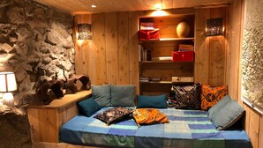 Une pièce chaleureuse avec un lit en bois, un mur en pierre et une étagère remplie de livres et surmontée d’une lampe.