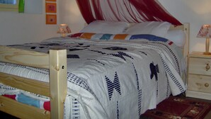 Ein Bett mit weiß-blauer Bettdecke, einem Holzkopfschild und einem roten Baldachin. Zwei Nachttische mit Lampen und ein kleines Wandbild befinden sich über dem Bett.