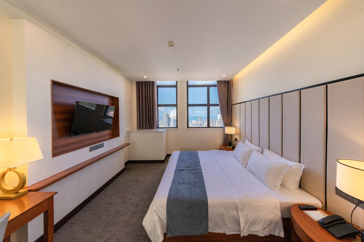 Ein Hotelzimmer mit Bett, Wandfernseher, Schreibtisch mit Lampe, Nachttisch mit Telefon und Blick auf die Stadt durch große Fenster.