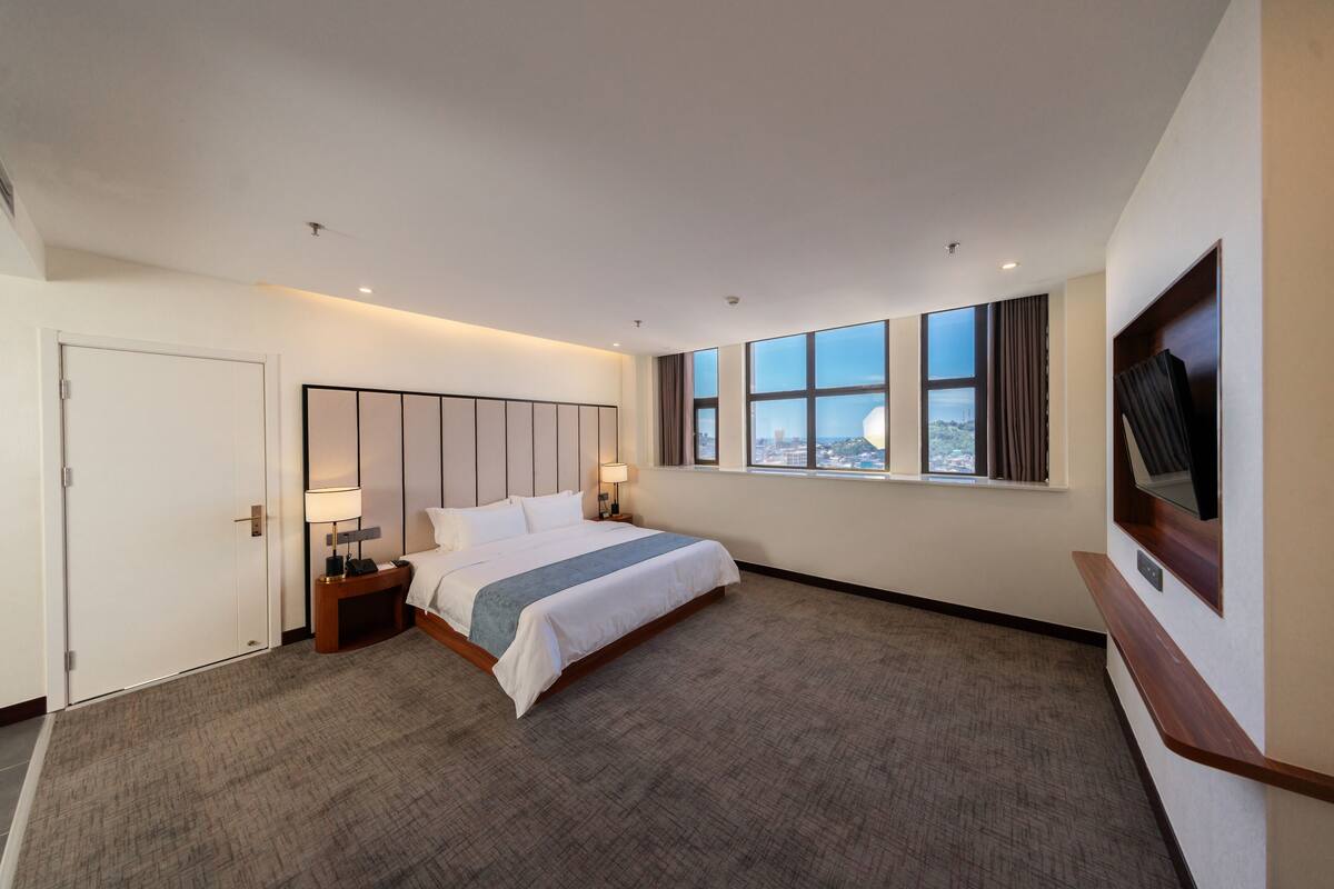 Ein Hotelzimmer mit einem großen Bett, einem Nachttisch, einem Fernseher und einem Fenster mit Blick auf die Stadt.
