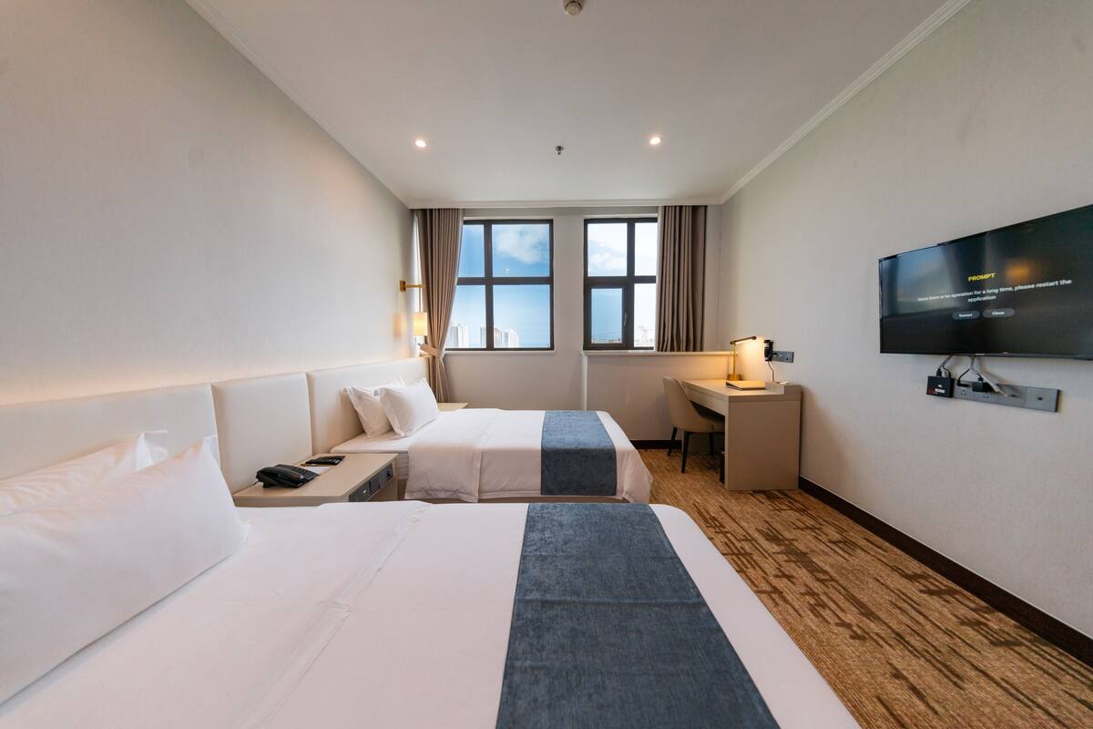 Ein Hotelzimmer mit zwei Betten, einem Schreibtisch, einem Fernseher und einem Fenster mit Blick auf die Stadt.