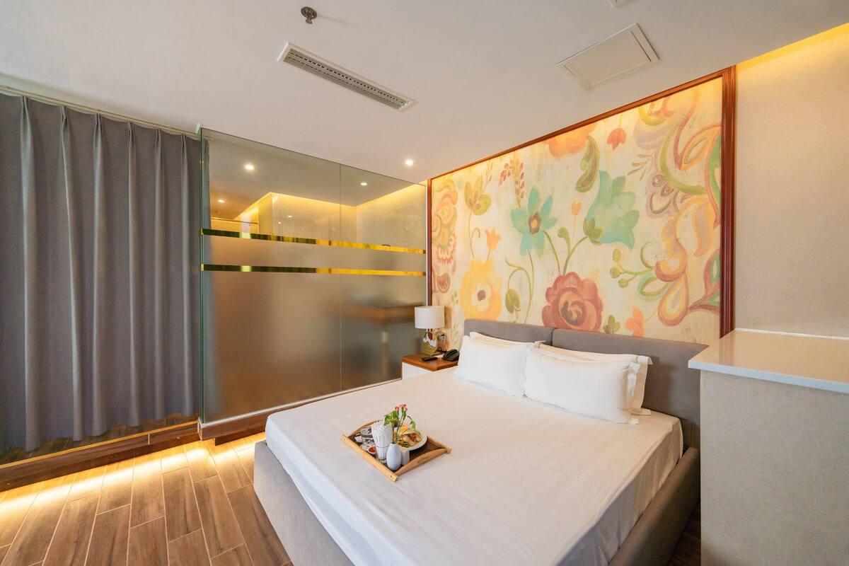 Ein modernes Hotelzimmer mit einem großen Bett, einer Glaskabine, floralem Wandbild und einem Tablett mit Frühstücksartikeln auf dem Bett.