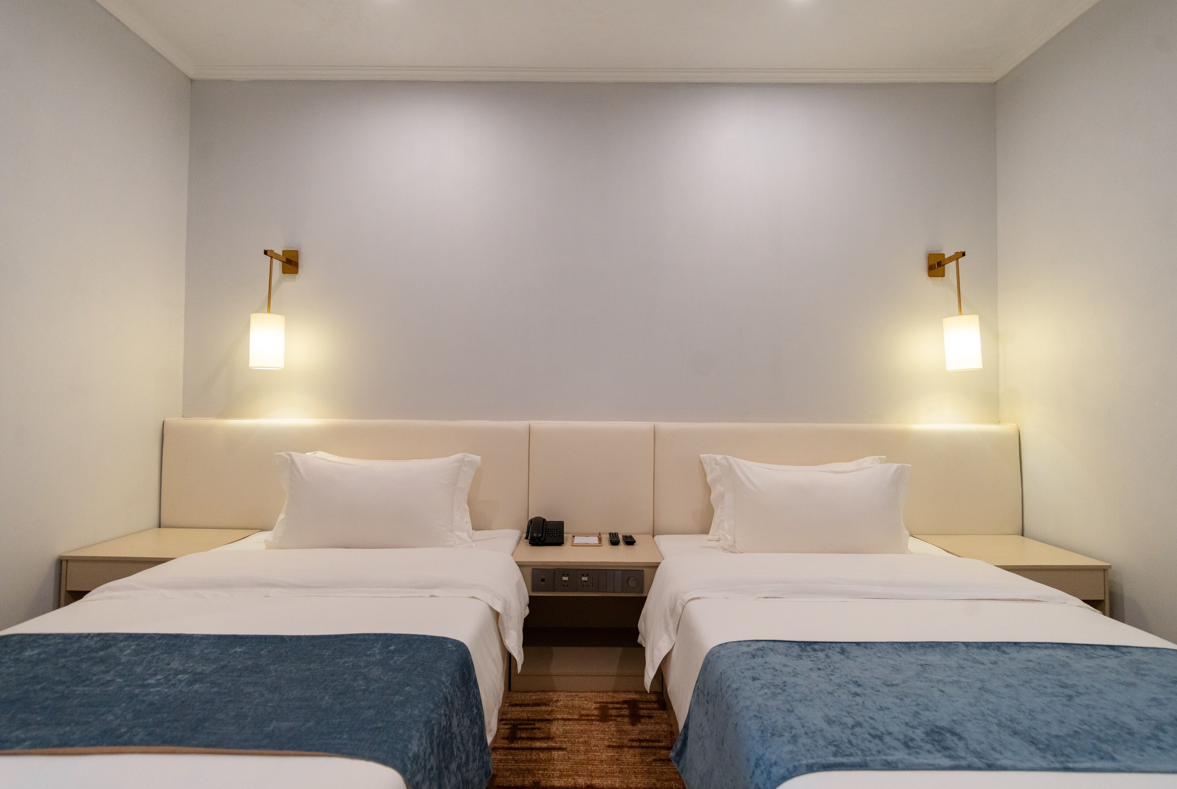 A hotel room with two beds, a nightstand, a desk, and two wall-mounted lamps.