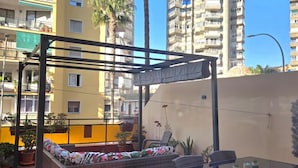 Un balcone con un divano in vimini, un tavolino in vetro e sedie. Sullo sfondo, piante in vaso e un lampione.