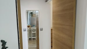 Un corridoio con una porta in legno e uno specchio che riflette una stanza con un lavandino e una pianta.