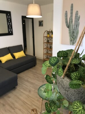 Ein Zimmer mit einer schwarzen Couch, einem gelben Kissen, einer hängenden Pflanze mit gefleckten Blättern und einem gerahmten Bild an der Wand.
