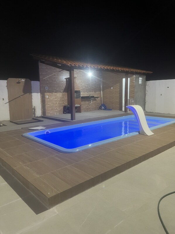 Ein Swimmingpool mit blauem Innenraum, umgeben von einer gefliesten Terrasse. Ein überdachter Bereich mit Mauerwerk und einer Tür ist vorhanden, und auf der Terrasse ist ein Schlauch sichtbar.