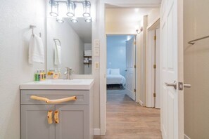 A bathroom with a sink, mirror, and toiletries. There is a hallway leading to a bedroom with a bed.