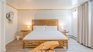 Un dormitorio con una cama de madera, dos mesitas de noche, una pequeña mesa auxiliar, un banco de madera y una lámpara de pared.
