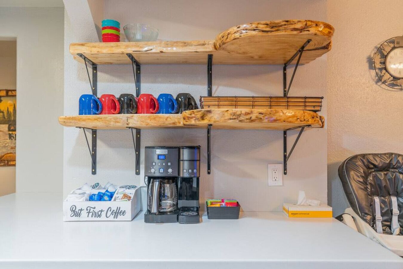 A kitchenette with a coffee maker, cups, and a box labeled "But First Coffee."