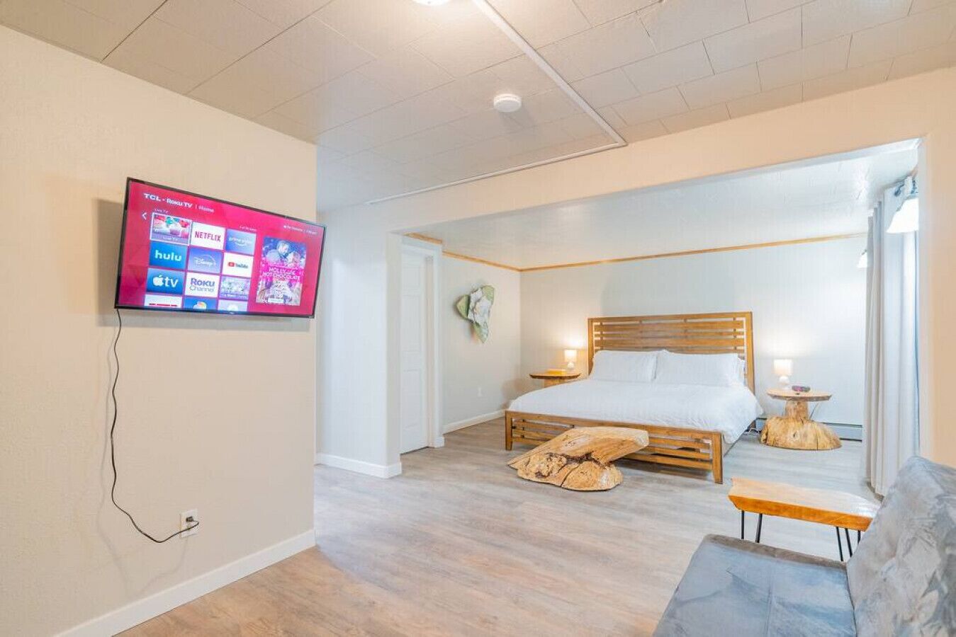 A modern hotel room with a wooden bed, a TV mounted on the wall, a small round table, and a bench.