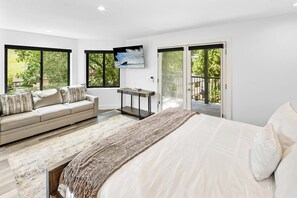 A modern bedroom with a large bed, a sofa, a TV, and a view of the outdoors through glass doors.