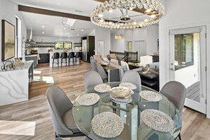 Un espace de vie moderne à aire ouverte, doté d’une table à manger en verre, de chaises recouvertes d’un tissu gris et d’un lustre décoratif.