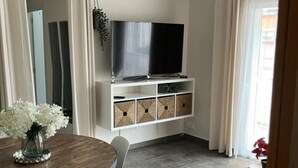 Un soggiorno moderno con una TV a schermo piatto montata su un mobile bianco, un tavolo da pranzo con sedie e un piccolo tavolino rotondo con una pianta.