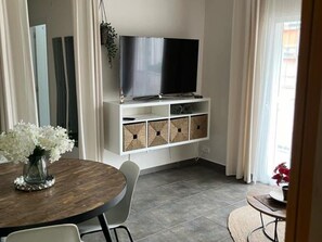 A modern living room with a flat-screen TV mounted on a white cabinet, a dining table with chairs, and a small round table with a plant. - Stunning 3BR Apartment in Torremolinos with Balcony Near Beaches and Plaza Andalucia (Torremolinos)