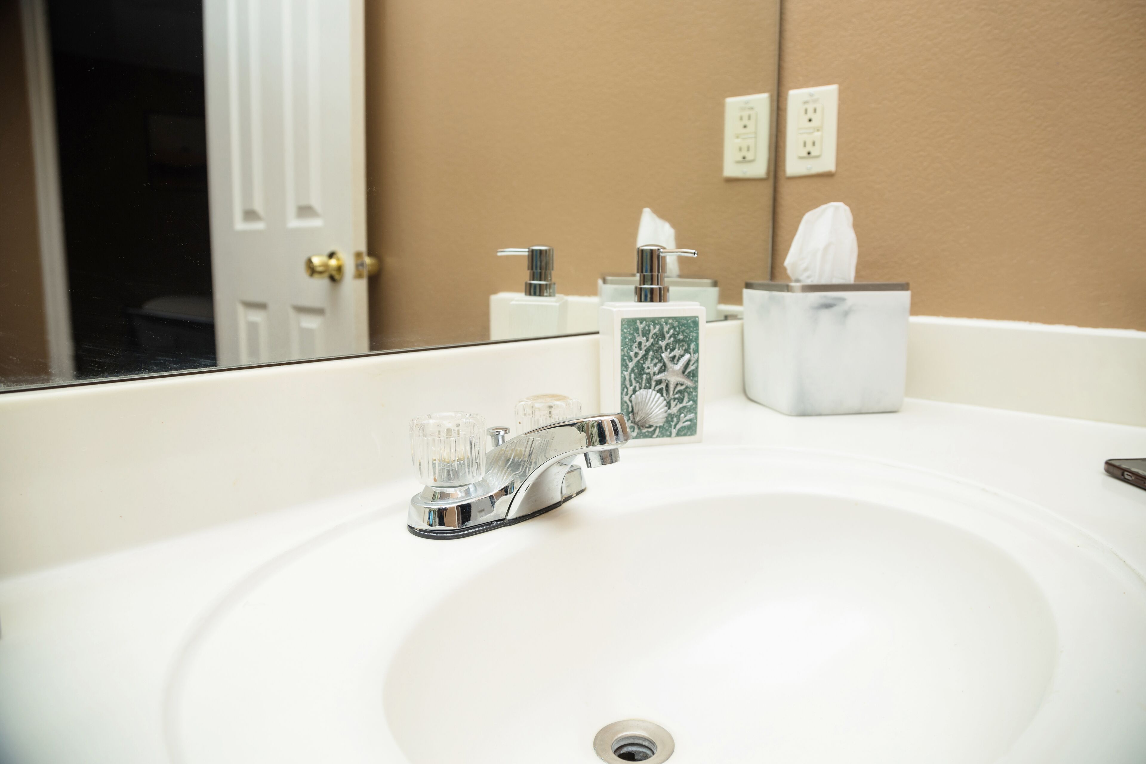 A bathroom with a white sink, chrome faucet, and a mirror reflecting a door and wall outlets.