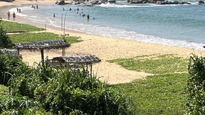 Ein Strand mit badenden und spazierenden Personen, einer felsigen Küstenlinie und einem kleinen Gebäude im Hintergrund.
