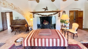 Un dormitorio amplio con una cama grande, vigas de madera en el techo, una chimenea, un piano y una silla.