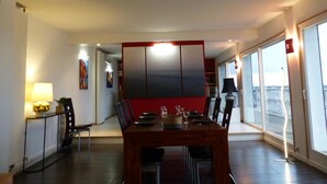 Un espace repas moderne avec une table en bois dressée pour quatre personnes, entourée de chaises. Un mur d’accent rouge, une grande fenêtre offrant une belle vue et une lampe sur la table d’appoint complètent l’ensemble.