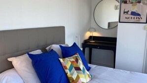 Un lit avec une literie blanche et plusieurs oreillers, une tête de lit rembourrée de couleur grise, une petite table de chevet avec une lampe et un miroir fixé au mur.