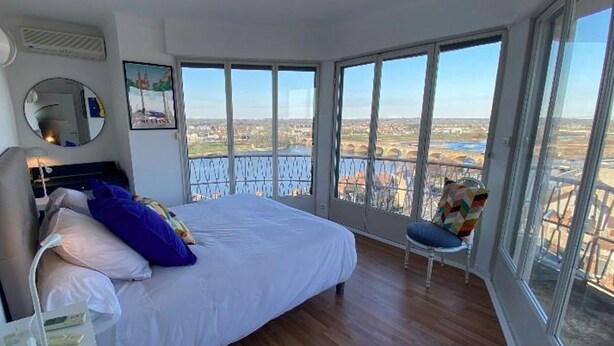 Une chambre avec un grand lit, une chaise et une vue sur le paysage urbain, visible à travers de grandes fenêtres.