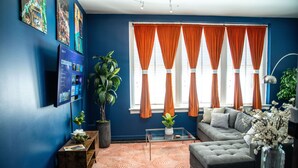 A modern living room with a blue accent wall, a large window with orange curtains, a grey sofa, and a glass coffee table. - Modern 1BR Downtown condo with rooftop gym & skyline views. King bed, Wi-Fi, walk to Canal & FQ (New Orleans)