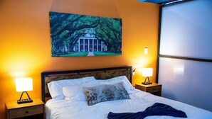 A neatly made bed with white linens and a blue throw blanket, flanked by two bedside tables with lamps, a large painting of a tree-lined building above the bed, and a window with blinds to the right. - Modern 1BR Downtown condo with rooftop gym & skyline views. King bed, Wi-Fi, walk to Canal & FQ (New Orleans)
