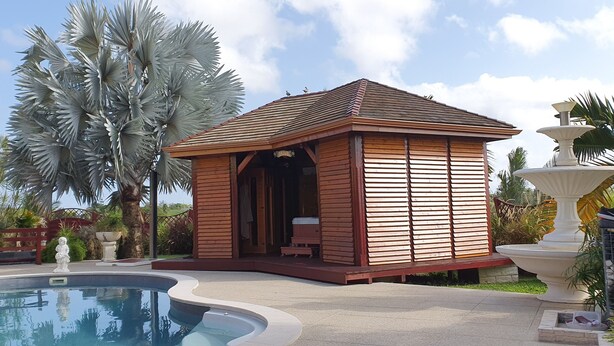 Un quiosco con techo y paredes de madera, una piscina y palmeras al fondo.