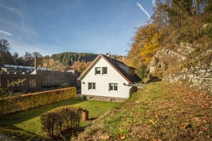 Una casa bianca con tetto spiovente, fondamenta in pietra e un piccolo giardino antistante con siepe e un albero.