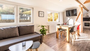 A modern living room with a sofa, dining table, and chairs, a TV mounted on the wall, and a view of the outdoors through large windows. - Zur Altstadt 3 - modern apartment just a few minutes' walk from the historic old town (Monschau)