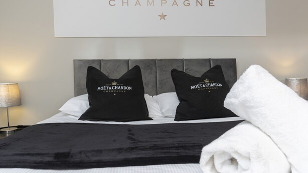 A hotel room with a bed featuring black and white bedding, two black pillows with 'MOËT & CHANDON CHAMPAGNE' branding, and a white towel. - Champagne House - 3 Bedroom Townhouse (Darlington)