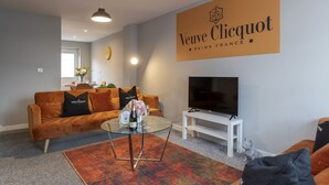 A living room with a sofa, a glass coffee table, a flat-screen TV, and a sign reading "Veuve Clicquot Reims France". - Champagne House - 3 Bedroom Townhouse (Darlington)