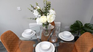 A dining table with a glass top surrounded by four brown upholstered chairs, white plates, and a vase with white flowers. - Champagne House - 3 Bedroom Townhouse (Darlington)