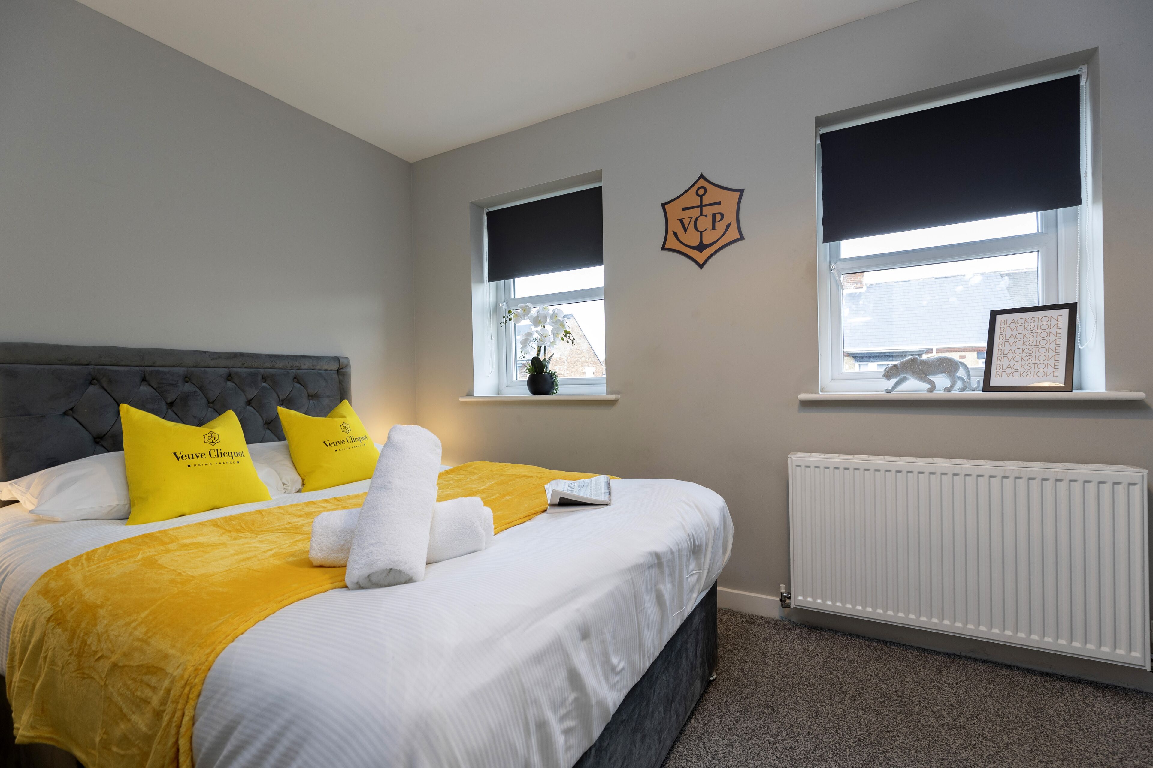 A neatly made bed with a grey headboard, white and yellow bedding, and a white towel. There are two windows with black blinds, one with a picture frame.